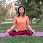 Woman meditating in lotus position on a yoga mat in a sunny park.