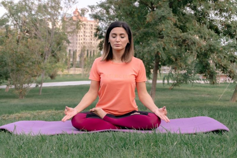 Woman meditating in lotus position on a yoga mat in a sunny park.
