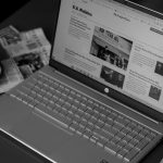 Black and white image of a laptop displaying news articles, accompanied by a cup of coffee and newspapers.